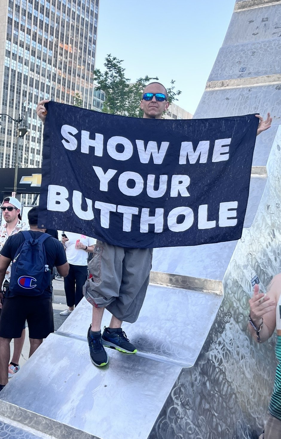 A man on a silver monument holding a black banner that reads 'show me your butthole', surrounded by a crowd in downtown Detroit.